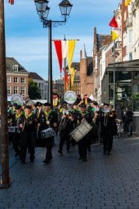 Straatparade met marching band in zwarte uniformen en gele sjaals; vlaggen langs historische straat.