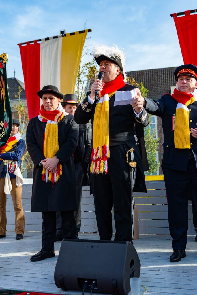 Podium met mannen in uniformen met gele en rode sjaals; man met pluimenhoed, microfoon en zwaard.
